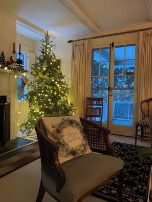 A living room with a Christmas tree, a fireplace, a large window with curtains, and a wooden chair with a cushion.