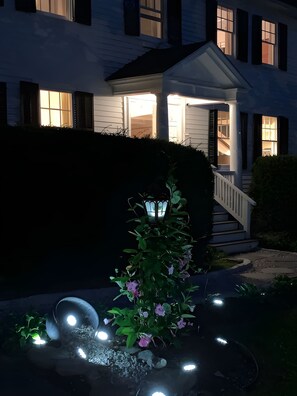 A residential house with a well-lit front porch and a garden with a small tree and lights.