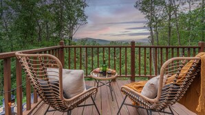 Une terrasse en bois, agrémentée de chaises en osier et d’une petite table, qui offre une vue sur une zone boisée et permet d’admirer le coucher du soleil.