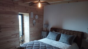A bedroom with a wooden wall, a bed with a quilted cover, two patterned pillows, a bedside table with a lamp, and a hanging light fixture.