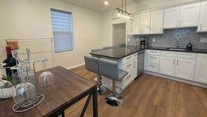 Una cocina moderna con armarios blancos, encimera negra y una zona de comedor con mesa de madera y dos taburetes de bar.