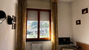 Una habitación con ventana, cortinas, un radiador, una repisa con un objeto pequeño y un cuadro colgado en la pared.