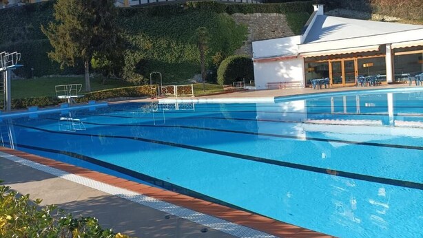 Una piscina con agua azul cristalina, rodeada por un solárium empedrado. Al fondo, un edificio con varios balcones y una zona cubierta con mesas y sillas.