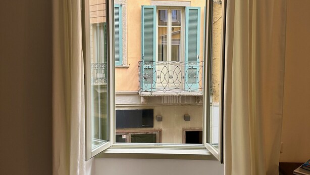Een raam met uitzicht op een gebouw met blauwe luiken en een balkon met een metalen hekwerk.