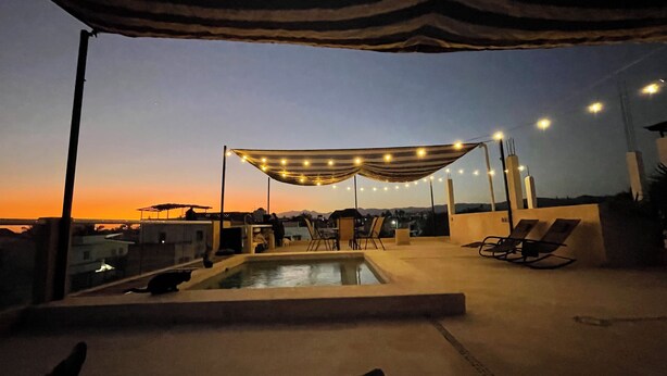 Zona de piscina en la azotea con piscina, sillones de descanso e incluso una pérgola con luces de cuerda.
