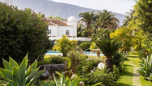 A lush garden with a pathway leading to a white building with a dome, surrounded by various plants and a mountain in the background.