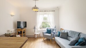 Ett modernt vardagsrum med ett soffbord i trä, en grå soffa med blå kuddar, ett litet vitt sidobord, en tv på ett träställ och ett fönster med tunna, genomskinliga gardiner.
