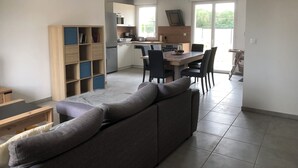 A modern living room with a grey sofa, a wooden bookshelf, and a dining area with a table and chairs. - Gîte les Coquelicots (Garennes-sur-Eure)