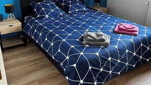 A bedroom with a blue wall, a bed with a geometric patterned bedspread, a framed photograph above the bed, and a wooden floor. - Gîte les Coquelicots (Garennes-sur-Eure)