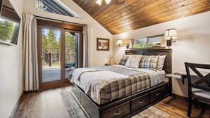 A bedroom with a wooden ceiling, a bed with a plaid bedspread, a desk with a lamp, and a view of the outdoors through a sliding door. - 2 Miles to Going-to-the-Sun Road - Glacier NP – 8 Private Wooded Acres – Hot Tub (West Glacier)