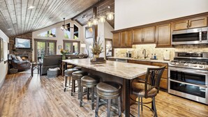 A spacious kitchen with wooden cabinets, a large island with stools, a built-in oven, and a stone fireplace. - 2 Miles to Going-to-the-Sun Road - Glacier NP – 8 Private Wooded Acres – Hot Tub (West Glacier)