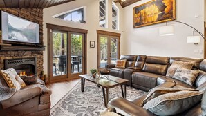 A spacious living room with a fireplace, leather sofa, and a large painting. - 2 Miles to Going-to-the-Sun Road - Glacier NP – 8 Private Wooded Acres – Hot Tub (West Glacier)