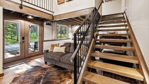 A spacious living area with a large window, a wooden staircase, and a cozy seating area. - 2 Miles to Going-to-the-Sun Road - Glacier NP – 8 Private Wooded Acres – Hot Tub (West Glacier)