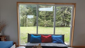 A room with a large window, a sofa with blue and red cushions, a wooden floor, and a small table with a vase and flowers. - Cabin near Frutillar Lake Llanquihue (Casma)
