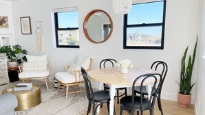 Un comedor con una mesa redonda, sillas negras y un mantel blanco. Hay dos ventanas con marcos negros, una pared blanca y una lámpara colgante.