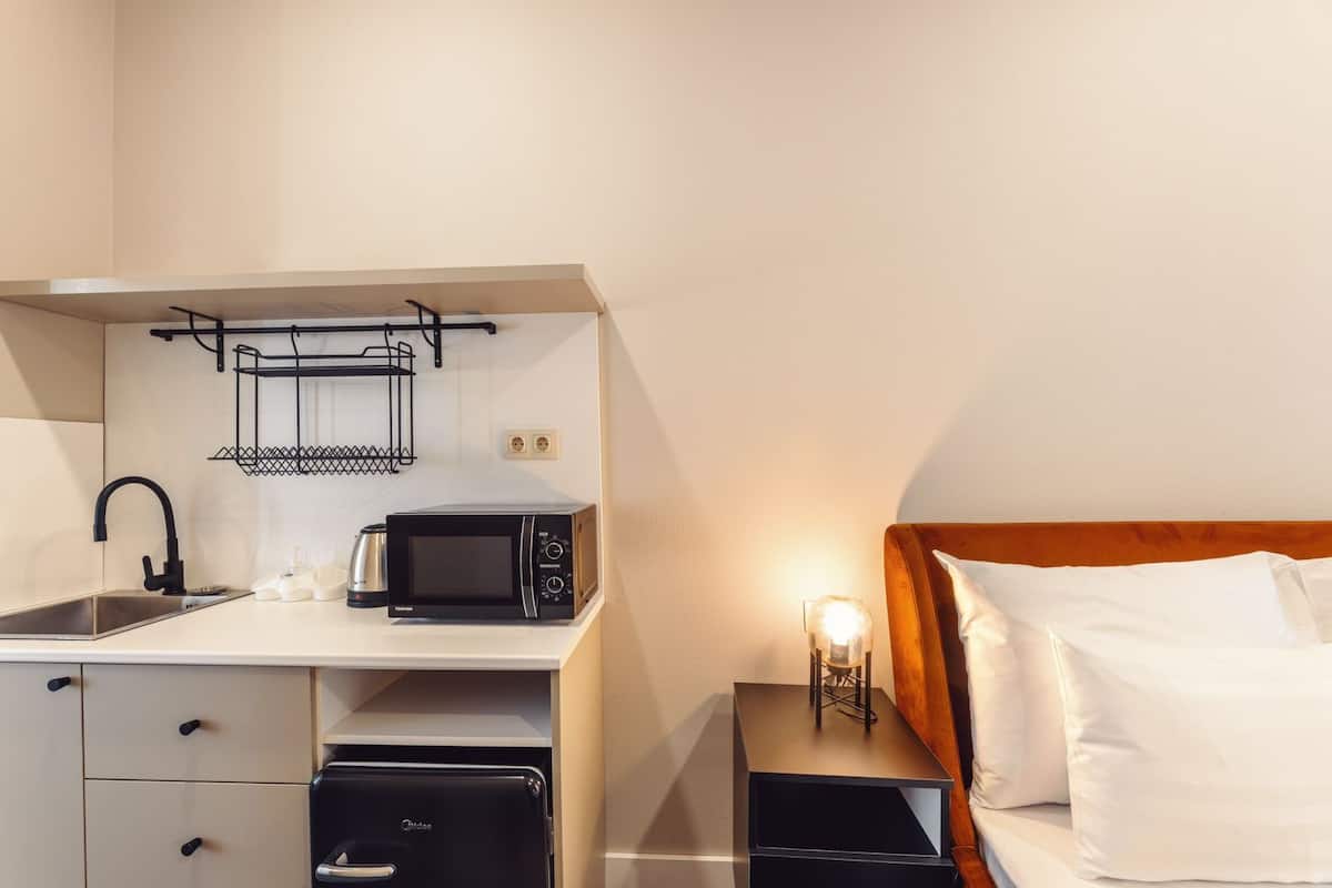 A compact kitchenette with a microwave, toaster, and sink. There is a bed with white pillows and a bedside table with a lamp.