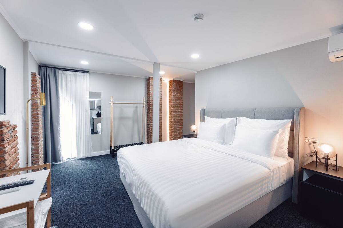 A modern hotel room with a large bed, a desk, a chair, a wardrobe, and a bathroom visible through an open door.