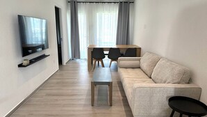 Ein modernes Wohnzimmer mit Flachbildfernseher an der Wand, einem beigefarbenen Sofa, einem Holztisch und einem Essbereich mit Tisch und Stühlen.