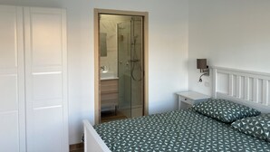 A bedroom with a bed, a bedside table, a nightstand, a hanging light fixture, a wardrobe, and a shower enclosure.