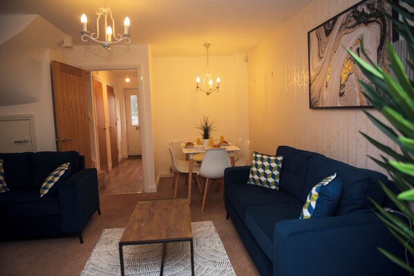 A living room with a dark blue sofa, a coffee table, and a chandelier. There is a dining area with a table and chairs. - Spacious 3 bed  town house in Bristol. (Bristol)