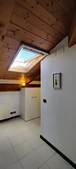 Una habitación con techo inclinado, tragaluz, armario blanco, radiador y un cuadro en la pared.
