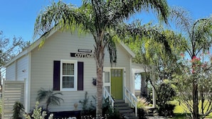 Ein weißes Cottage mit grüner Tür, beschriftet mit "COMMISSARY COTTAGE", mit Veranda und Kiesweg.