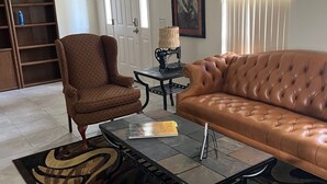 Un salon avec un canapé en cuir marron, un fauteuil à motifs, une table basse en verre, un ventilateur de plafond, une bibliothèque et un tableau encadré accroché au mur.