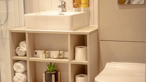 Un baño con lavamanos blanco, repisas con toallas y artículos de aseo, y un inodoro.