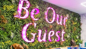 Una pared vibrante con un letrero que dice 'Be Our Guest', rodeada de exuberante follaje.