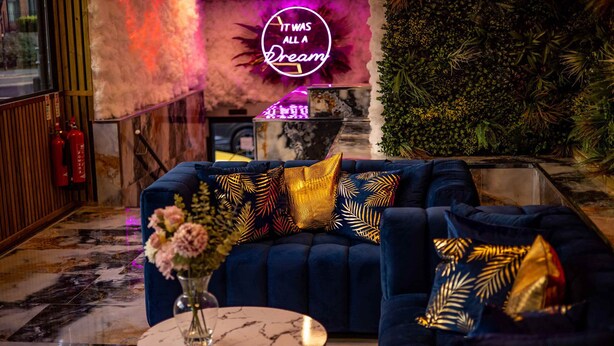 Un rincón lounge acogedor con un sofá azul oscuro, almohadones decorativos, una mesa de centro de mármol y un letrero de neón.