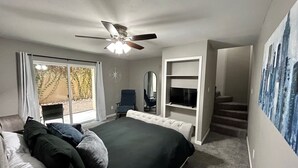 Un dormitorio con una cama grande, un ventilador de techo, un televisor, una silla y un ventanal corredizo a un balcón.