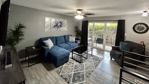 Una sala de estar con un sofá azul, una mesa de centro, un televisor y una zona de comedor con una silla. Hay un ventilador de techo, un reloj y vistas a través de puertas de vidrio.