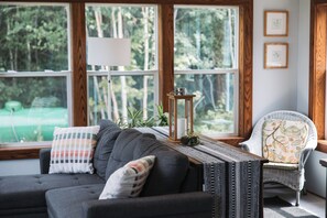 A cozy living room with a dark grey sofa, a patterned pillow, a wicker chair, and a view of a forest through large windows.