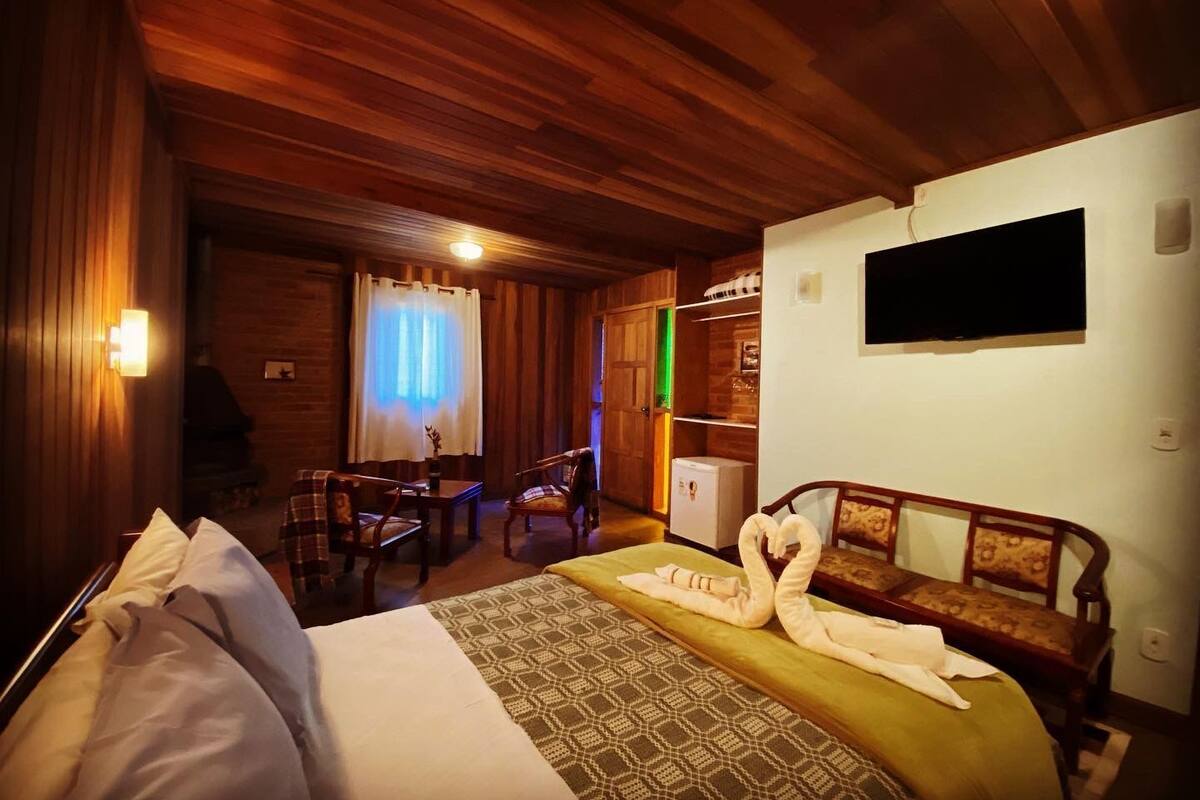 A cozy room with a bed, a wooden ceiling, a TV mounted on the wall, and a dining area with chairs and a table.