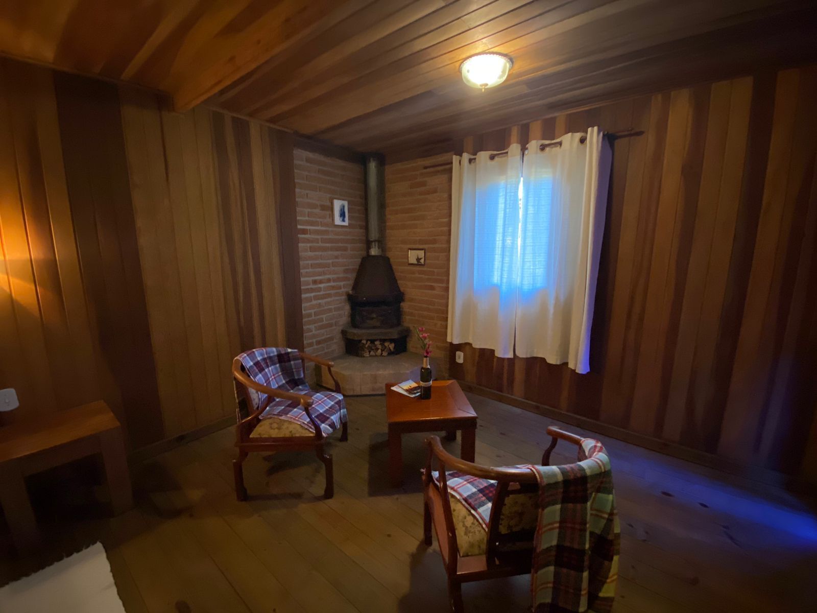 A cozy room with wooden walls and a brick fireplace. There are two plaid chairs and a small table.