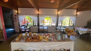A breakfast buffet with various pastries, fruits, and beverages on a table in a room with wooden ceilings and large windows. - Pousada Sotaque Mineiro (Cunha)