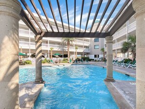 A pool area with a pergola overhead, lounge chairs, and a multi-story building in the background. - Beautiful Galveston, ocean front, Ocean views, Comfortable Beds N Amazing pools. (Galveston)