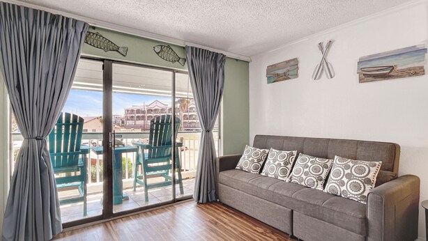 Quarto com sofá cinzento, almofadas estampadas, porta de vidro deslizante para área externa com cadeiras e vista de edifícios.