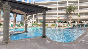 Área de piscina com espreguiçadeiras e um espaço de estar coberto, cercada por palmeiras e um edifício de vários andares ao fundo.