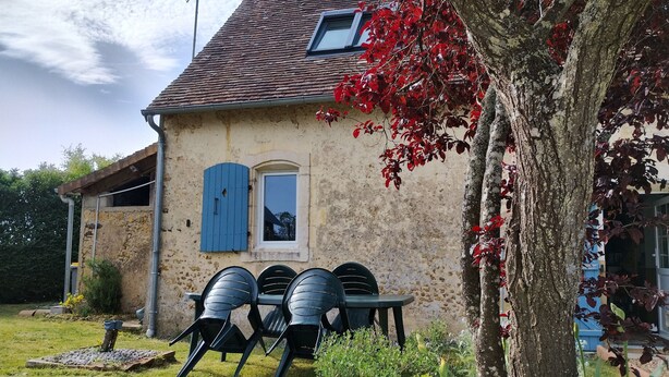 Una casa de piedra rústica con tejado de baldosas, una ventana pequeña con persianas azules y un árbol de hojas rojas al frente.