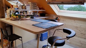 Una cocina con una mesa de madera, dos taburetes de bar, una ventana que ofrece vistas a árboles y varios utensilios de cocina en estantes.