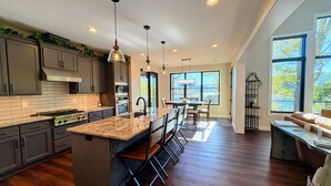 A spacious kitchen with a central island, dark wood flooring, and large windows offering views of the outdoors. - Norris Lakefront Retreat—Family Ready, Private Dock, Fire Pit, Screened Porch (LaFollette)