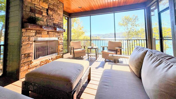 A covered outdoor seating area with a stone fireplace, wicker furniture, and a view of the lake. - Norris Lakefront Retreat—Family Ready, Private Dock, Fire Pit, Screened Porch (LaFollette)
