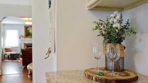 Una habitación con piso de madera, una puerta con picaporte dorado, un jarrón con follaje verde y flores blancas, y dos copas de vino vacías sobre un posavasos de madera.