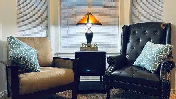 Una sala de estar con un sillón de cuero marrón y otro sillón de cuero negro con respaldo capitonado, ambos con cojines estampados. Una mesita con una lámpara y un objeto decorativo se encuentran entre ellos.