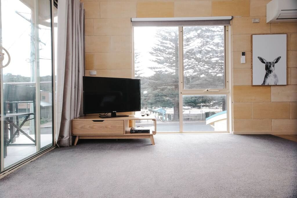 Un salon moderne avec un téléviseur à écran plat posé sur une console en bois, une porte-fenêtre coulissante donnant sur un balcon et un tableau encadré accroché au mur.