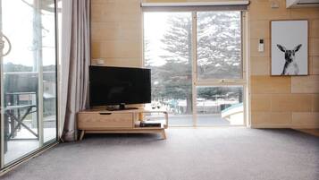 Un salon moderne avec un téléviseur à écran plat posé sur une console en bois, une porte-fenêtre coulissante donnant sur un balcon et un tableau encadré accroché au mur.