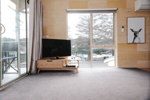 A modern living room with a flat-screen TV on a wooden console, a sliding glass door leading to a balcony, and a framed picture on the wall. - Surf break - Beachfront Port Campbell (Port Campbell)