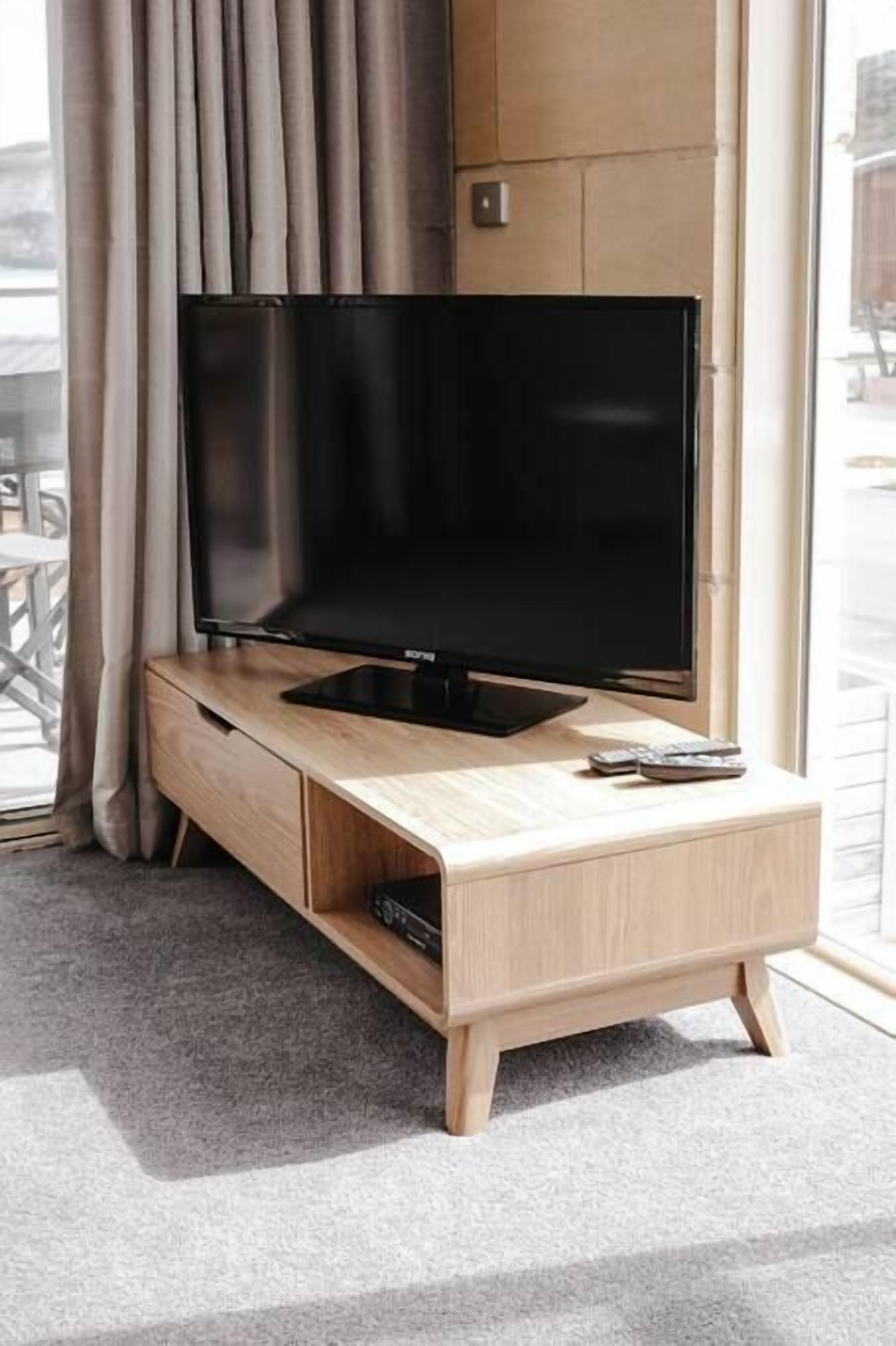 Een moderne woonkamer met een flatscreen televisie op een houten standaard, een afstandsbediening en een raam met gordijnen.