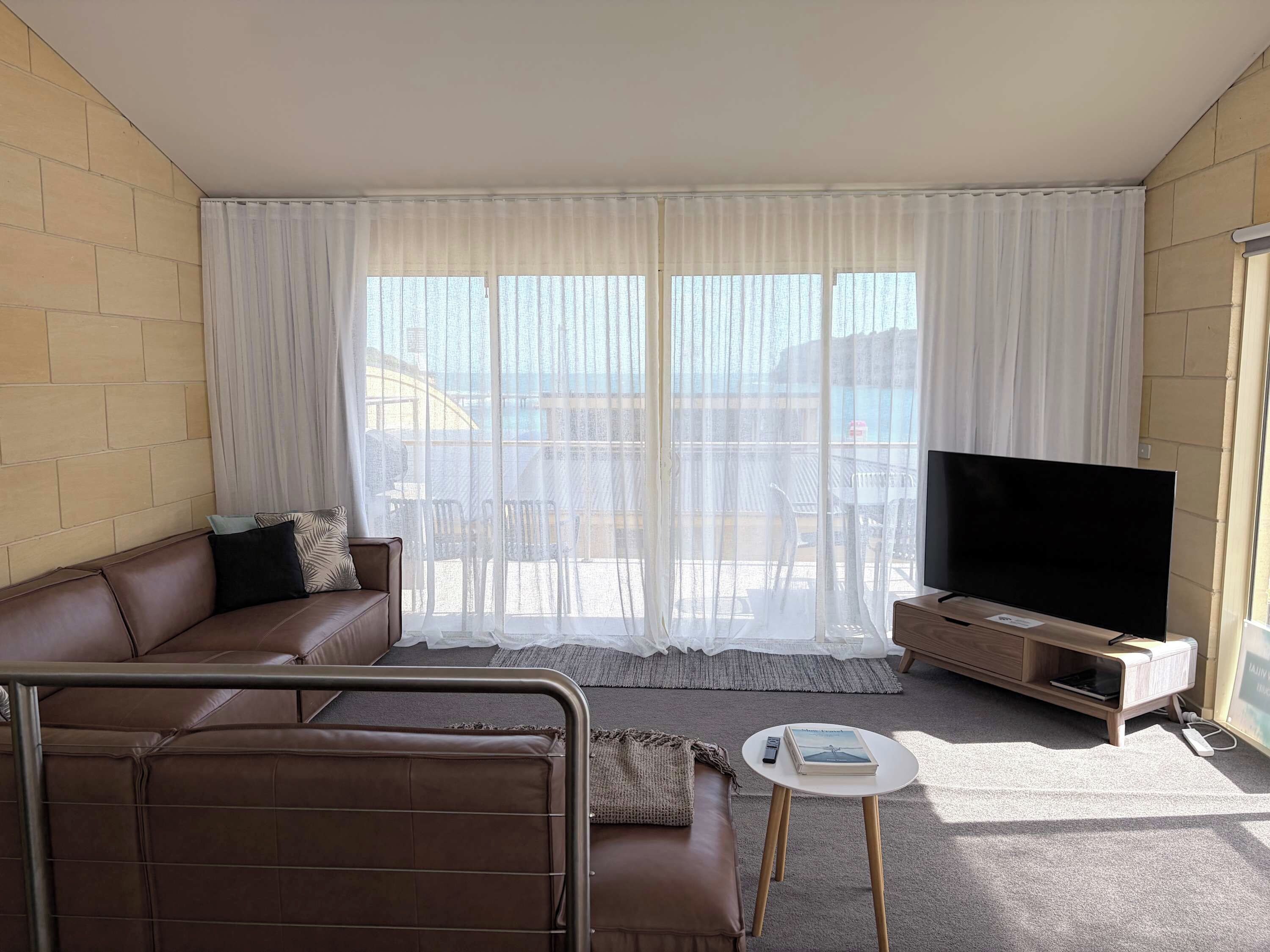 A living room with a brown leather sofa, a flat-screen TV on a wooden stand, and a large window with sheer curtains.